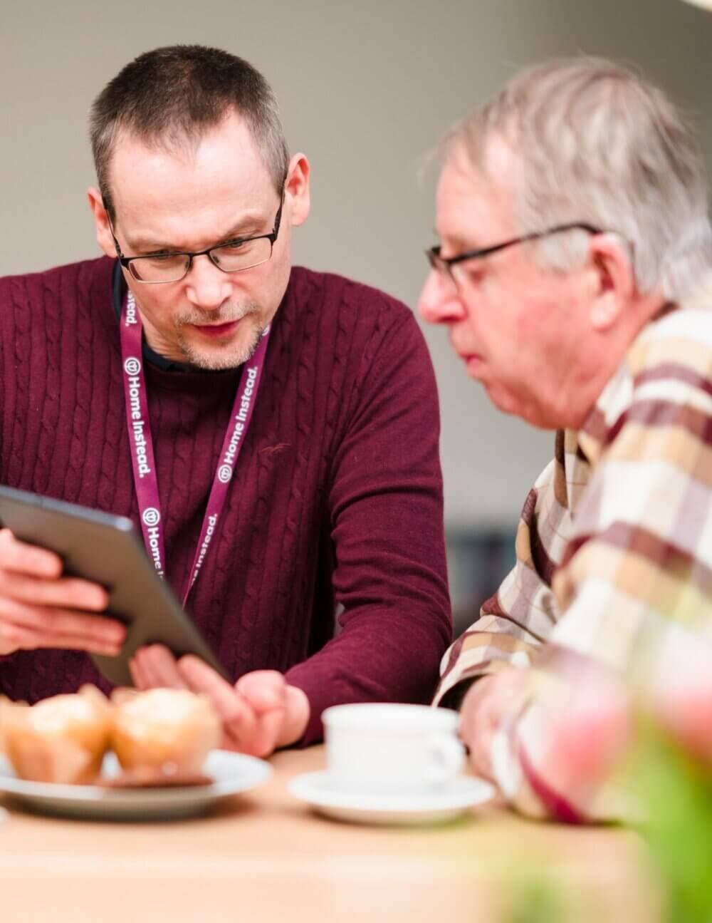 A man showing a tablet to an older man at a table with muffins and a cup. - Home Instead