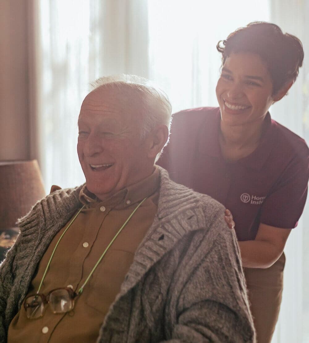 An elderly man in a sweater laughs with a caregiver smiling behind him, in a softly lit room. - Home Instead