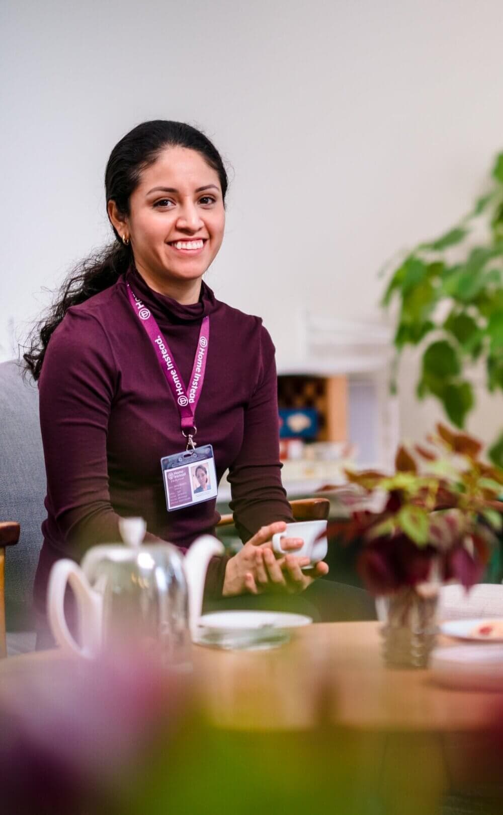 A smiling person with a lanyard and ID badge holds a cup of tea, sitting in a cozy room with plants and a teapot. - Home Instead