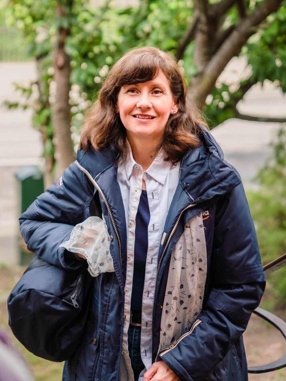 A woman wearing a dark jacket and carrying a bag smiles while standing outside with greenery in the background. - Home Instead