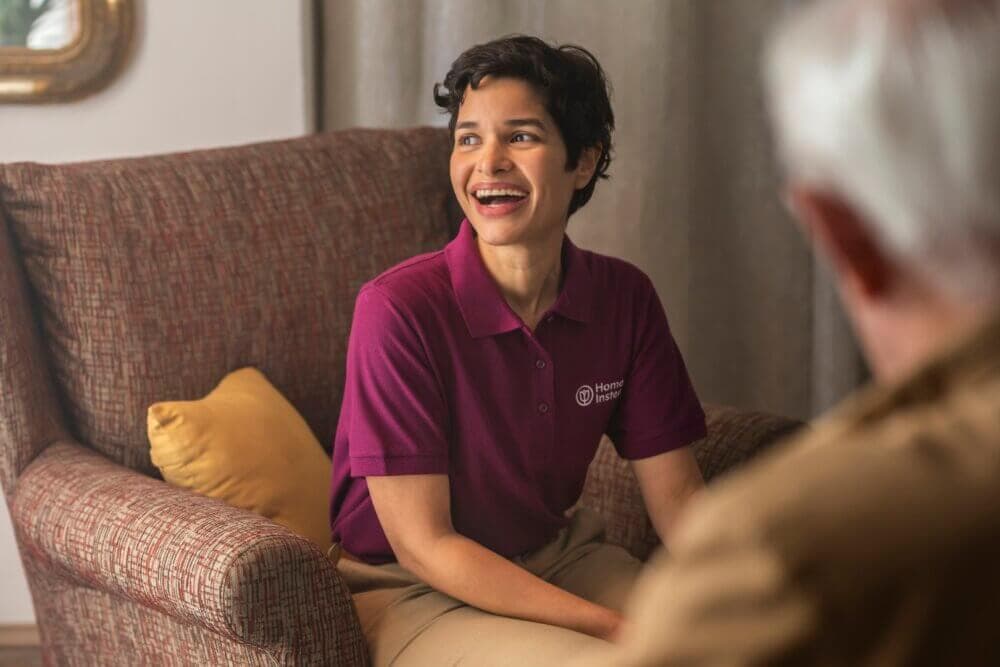 A woman in a maroon polo shirt sits on a chair, smiling at an older man whose back faces the camera. - Home Instead