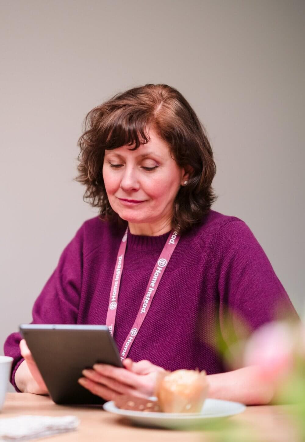 Woman with short brown hair, wearing a purple shirt and lanyard, reading a tablet at a table with a plate and cup. - Home Instead