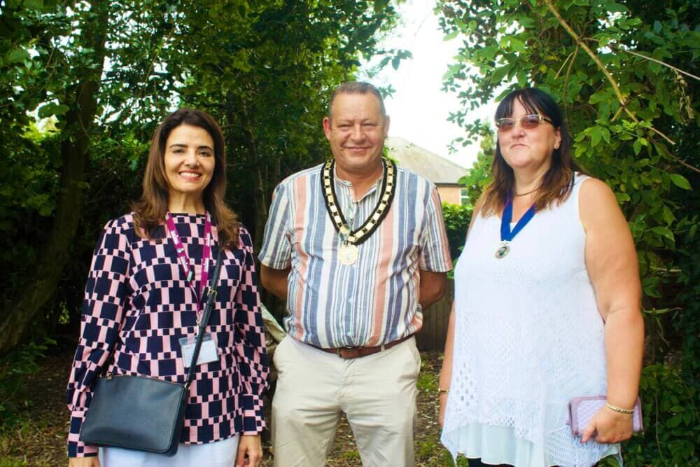 Three people standing together, smiling, with greenery in the background; a man in the center wearing a ceremonial chain. - Home Instead