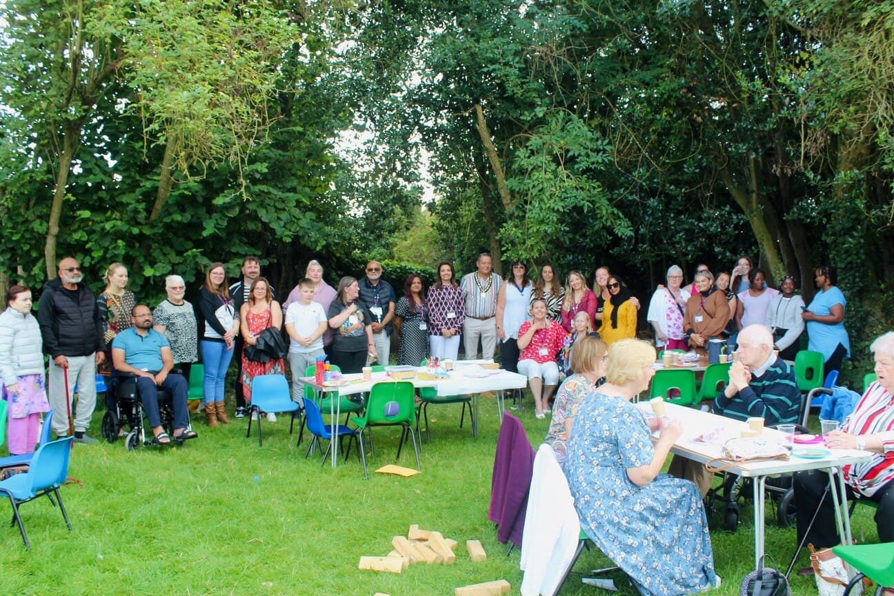 A diverse group of people gathers outdoors by trees, with tables and chairs for a communal event. - Home Instead