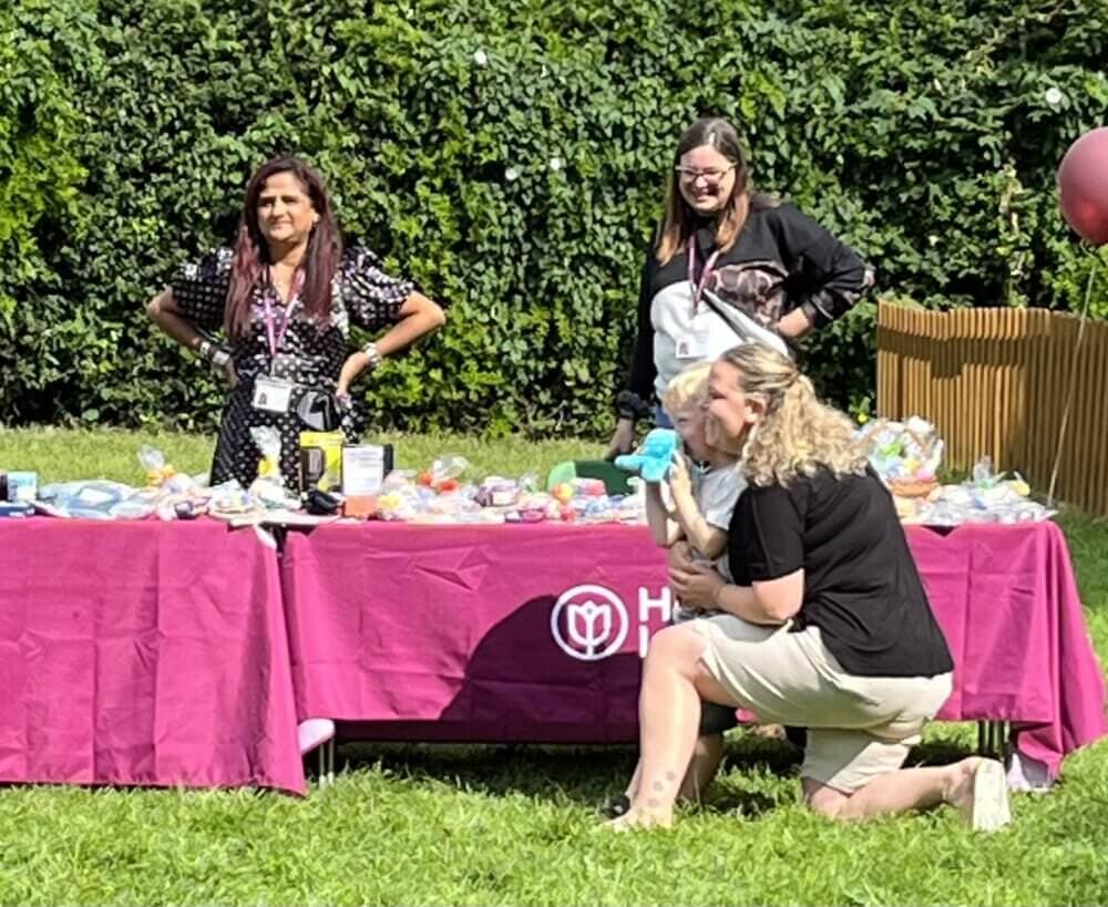 Three people stand behind a table with various items at an outdoor event. The table has a pink tablecloth. - Home Instead