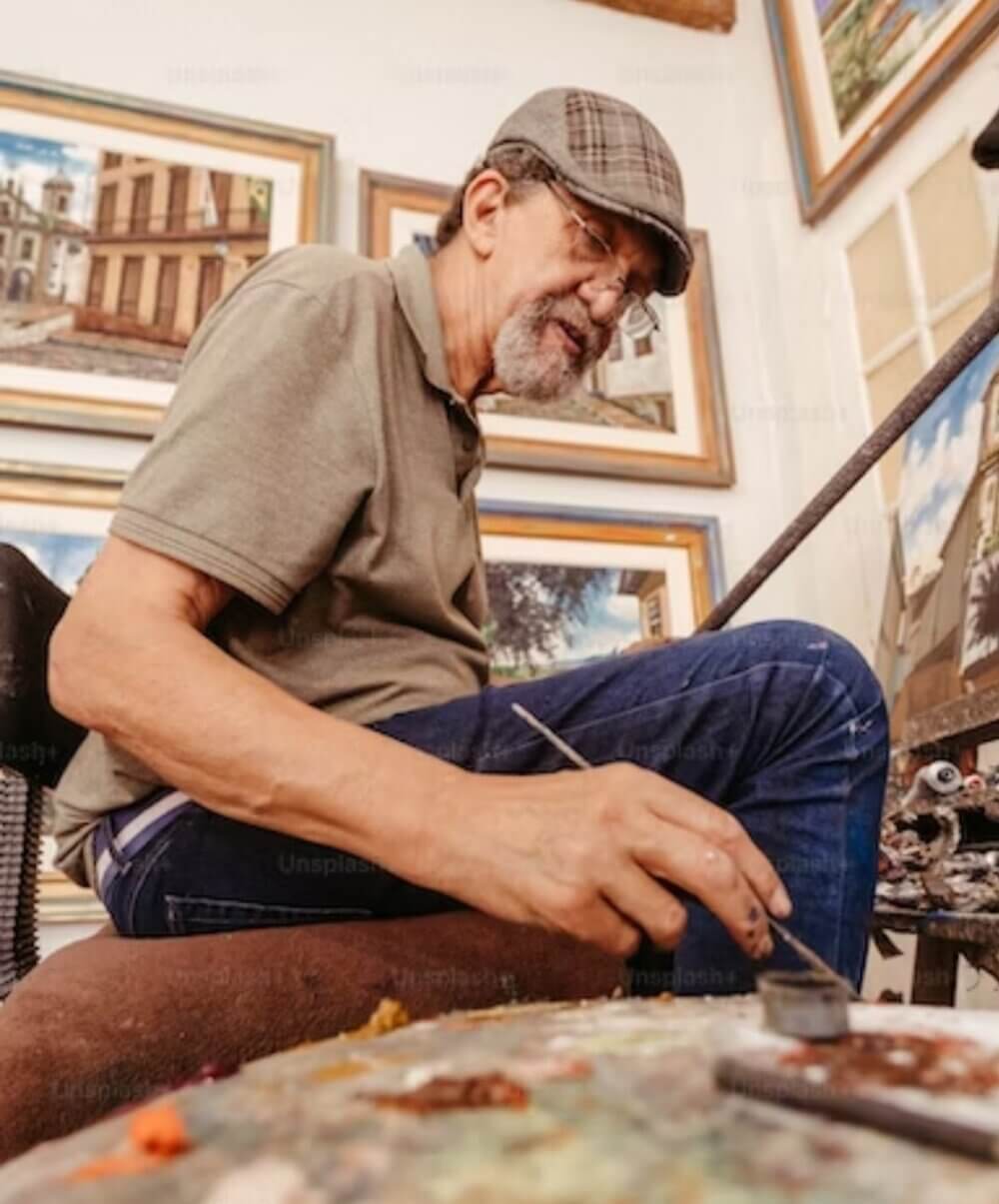 An older man wearing a cap is painting in an art studio surrounded by framed artworks. - Home Instead