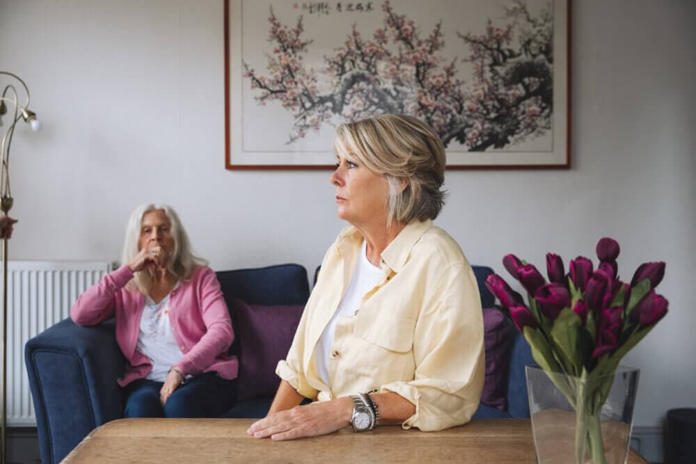 Two elderly women in a living room; one sits on a couch, the other at a table with a vase of tulips, both appearing pensive. - Home Instead