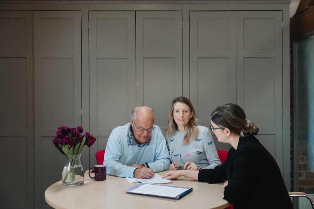 Three people sit at a table; one signs a document, while the others observe. A vase with flowers is in the foreground. - Home Instead