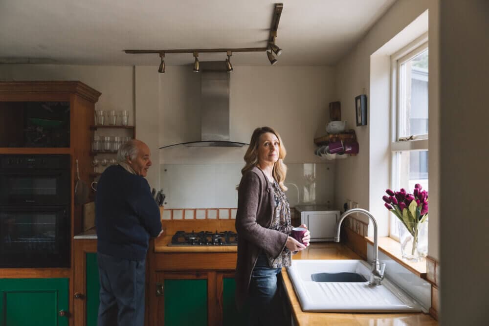 Two people in a kitchen, one older man near the stove, and a woman holding a mug by the sink with flowers on the windowsill. - Home Instead