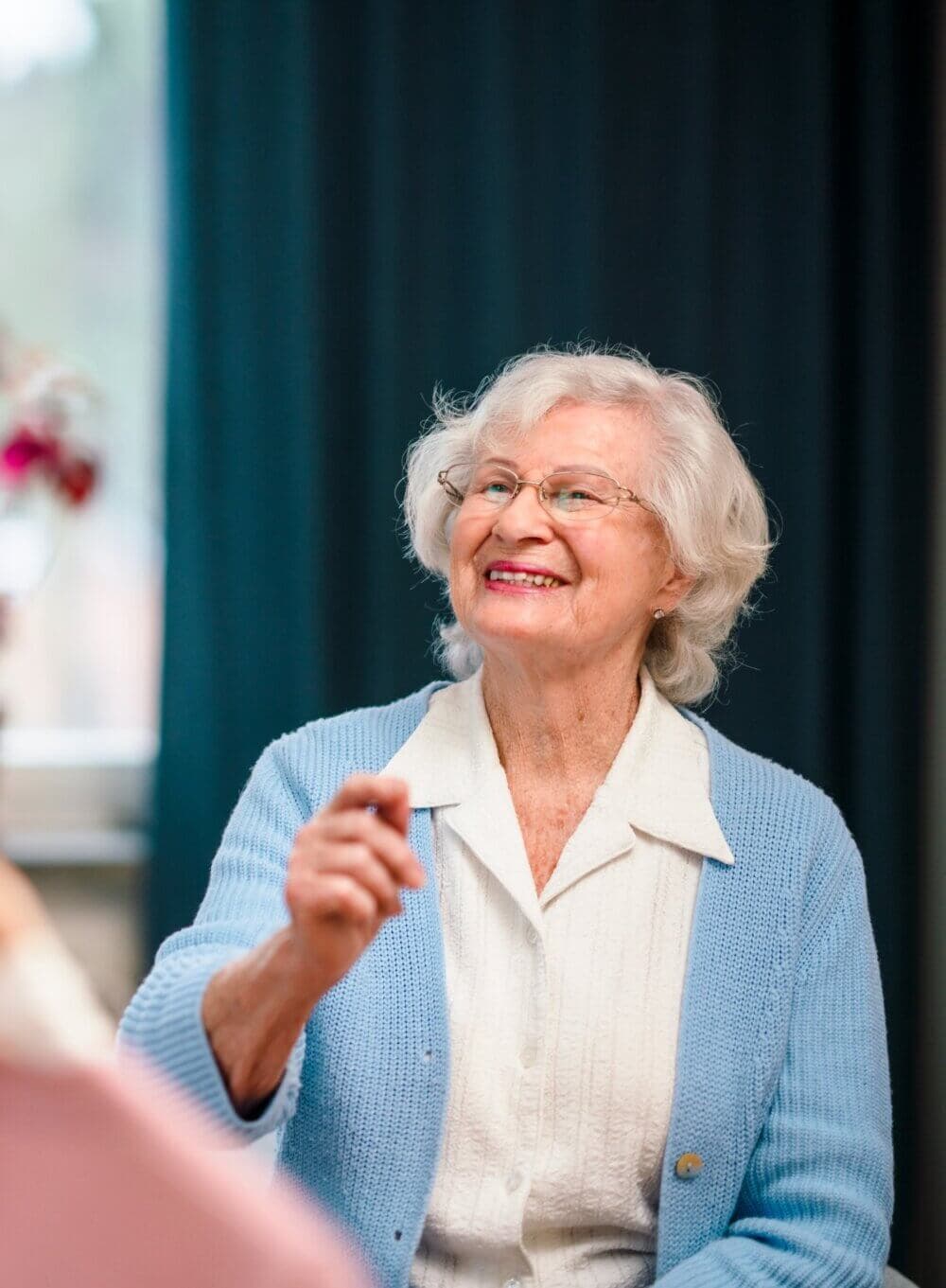 Smiling elderly woman with white hair and glasses wearing a white shirt and blue cardigan gestures with her hand. - Home Instead