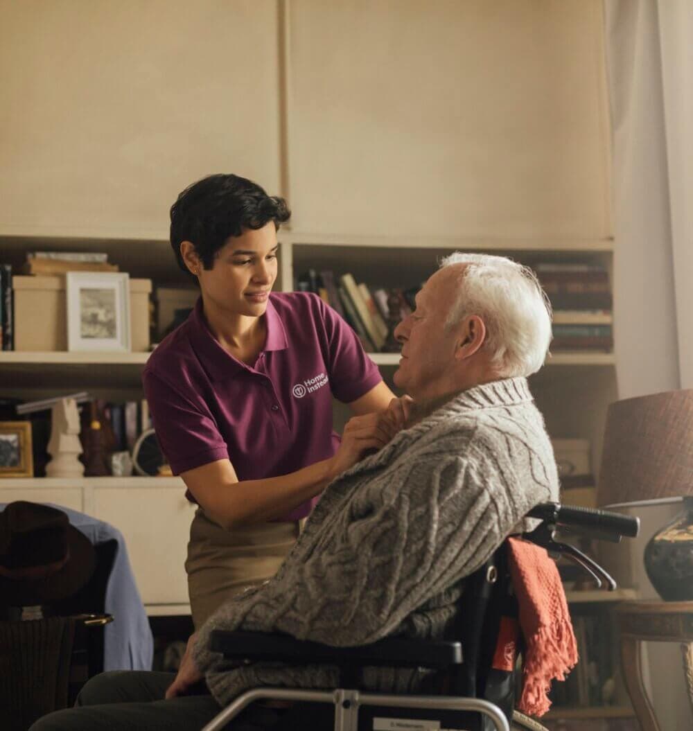 A caregiver in a purple shirt assisting an elderly man in a wheelchair inside a cozy, well-lit room. - Home Instead
