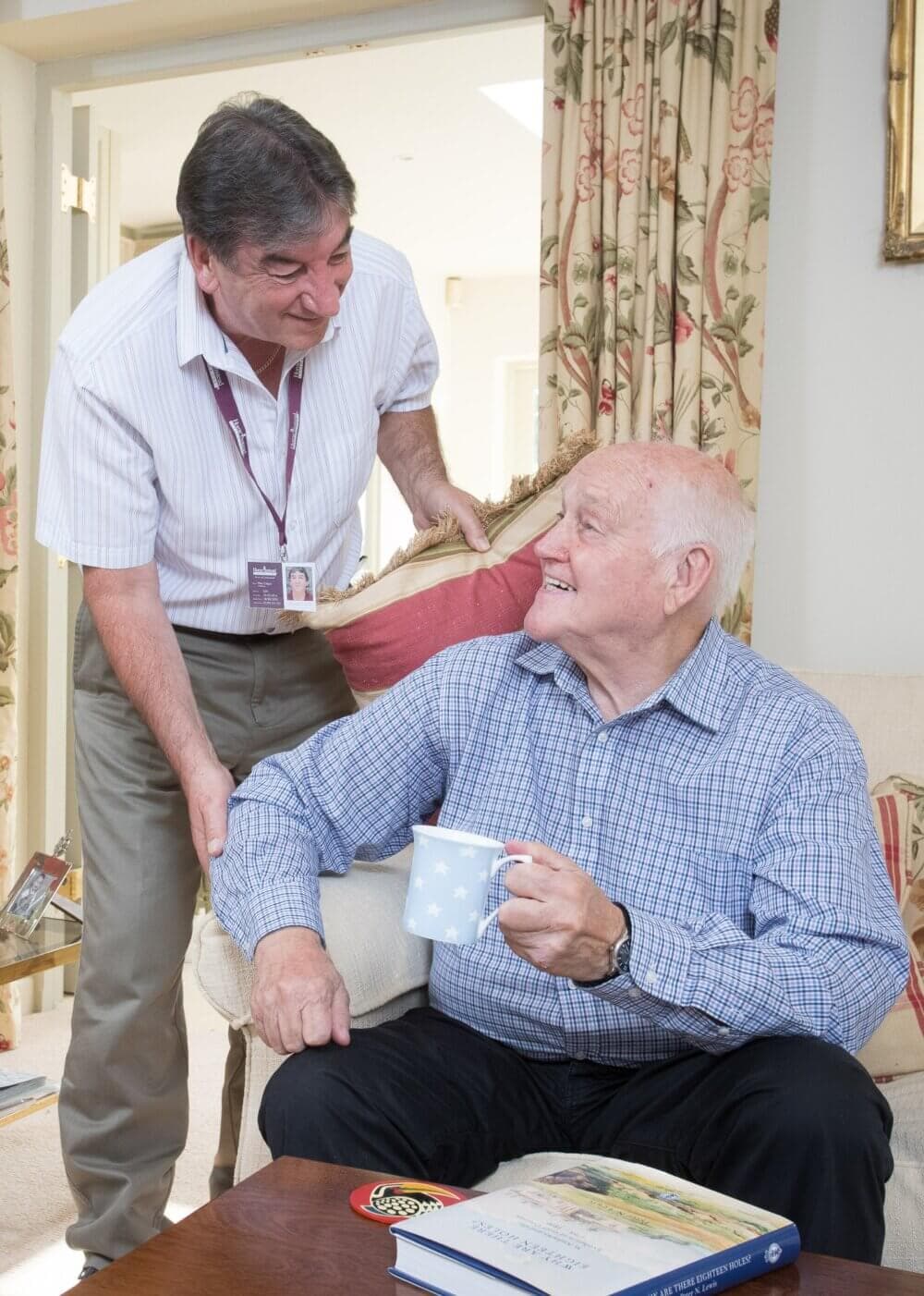 An elderly man holding a mug sits smiling and talking to a standing caregiver in a home setting. - Home Instead