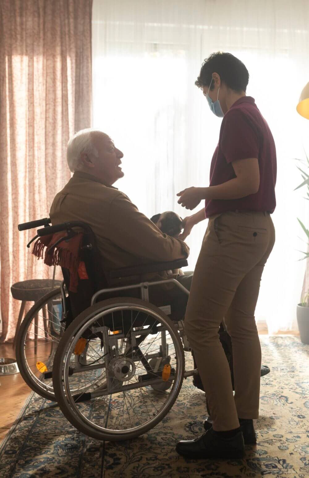 Elderly man in a wheelchair smiles while holding a small dog, as a standing caregiver in a mask assists him indoors. - Home Instead
