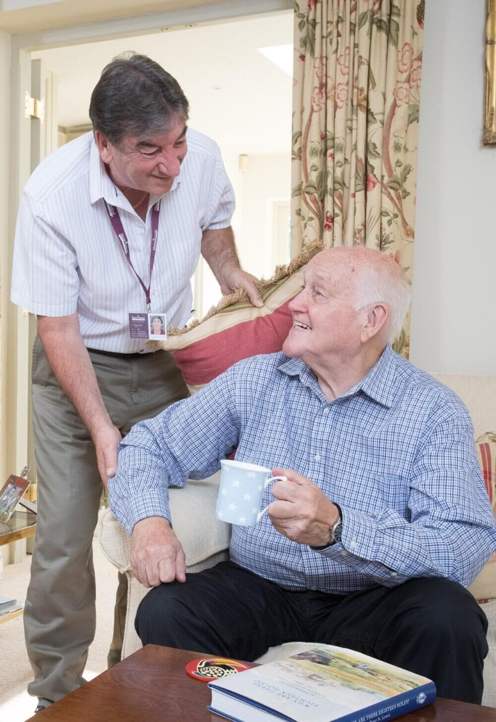 A caregiver assists a smiling elderly man holding a mug in a cozy room with floral curtains. - Home Instead