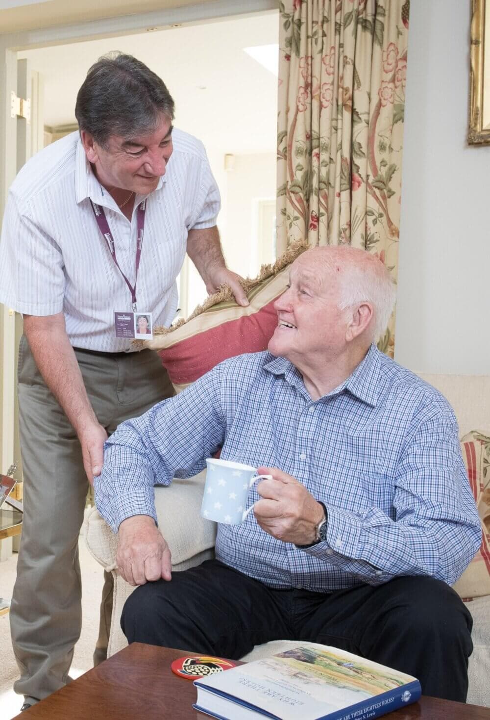 A senior man holding a mug is smiling at his caregiver, who is gently touching his arm in a cozy living room. - Home Instead
