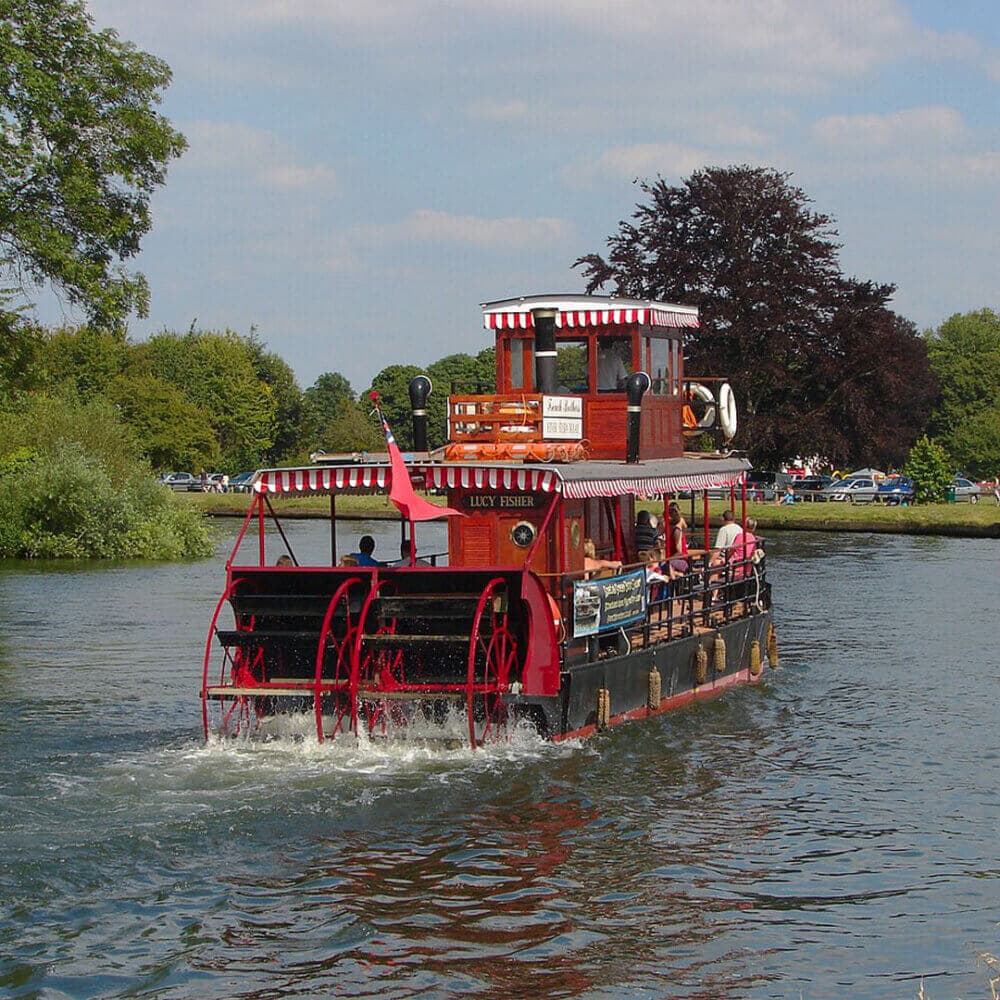 Red and white paddle steamer with passengers cruising on a river under a partly cloudy sky. - Home Instead