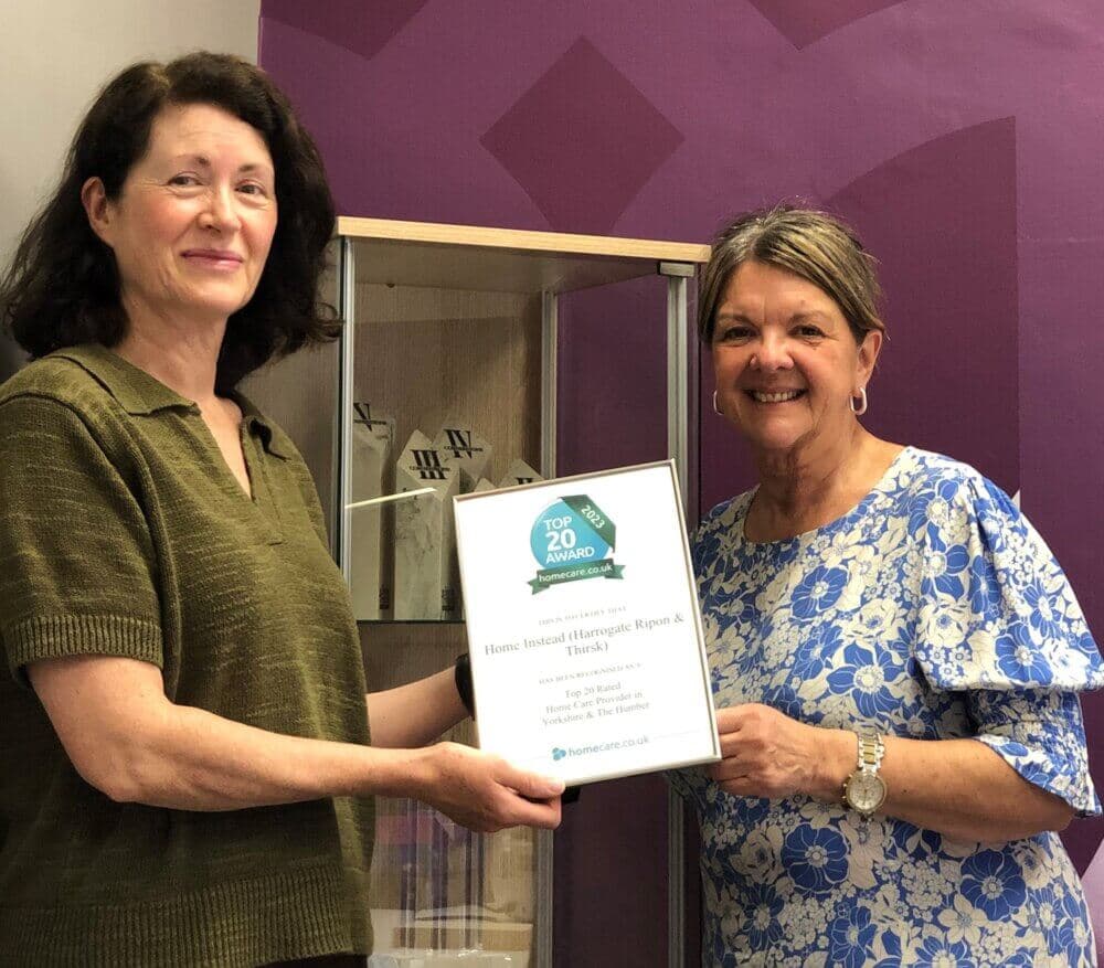 Two women smiling and holding a framed "Top 20 Award" certificate in a room with a purple wall and display case. - Home Instead