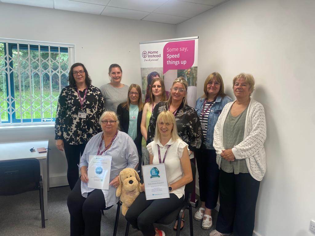 A group of people posing together in a room, two seated holding certificates, a stuffed toy in front. - Home Instead