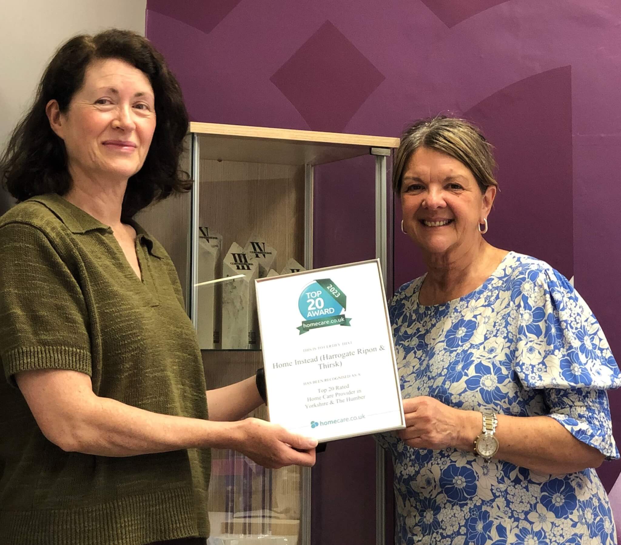 Two women smiling and holding a framed "Top 20 Award" certificate in a room with a purple wall and display case. - Home Instead