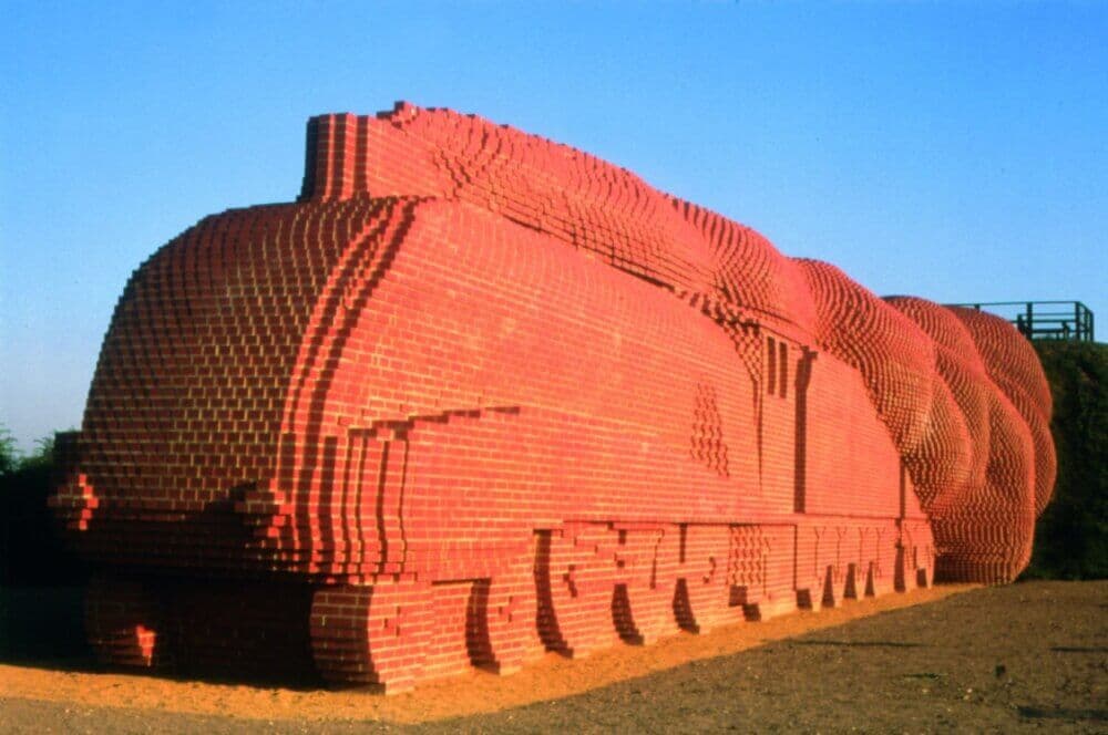 A large sculpture of a train made of red bricks, resembling a steam locomotive, set outdoors against a clear blue sky. - Home Instead