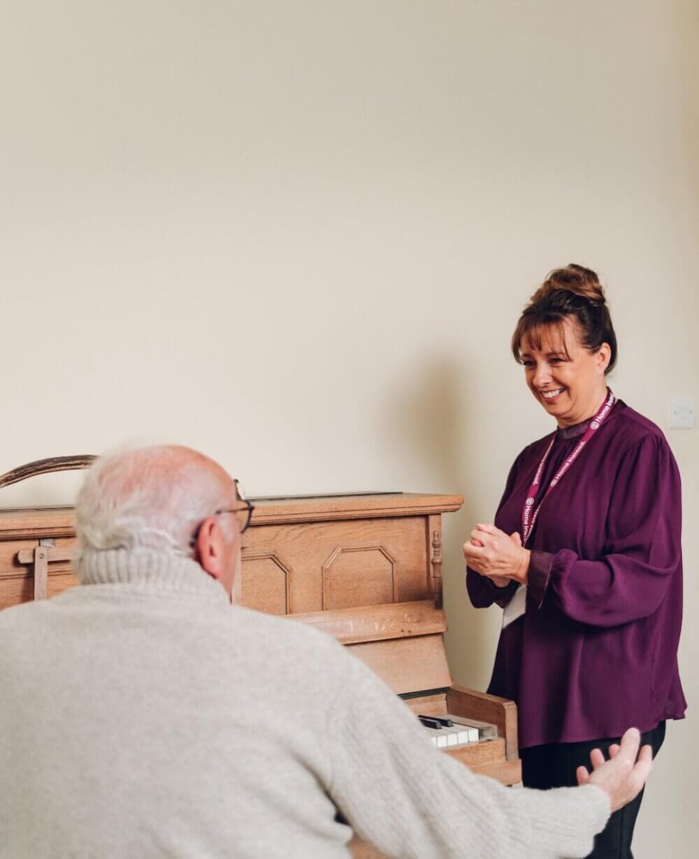 An elderly man plays the piano while a woman in a purple blouse watches and smiles in a warmly lit room. - Home Instead Bournemouth & Christchurch