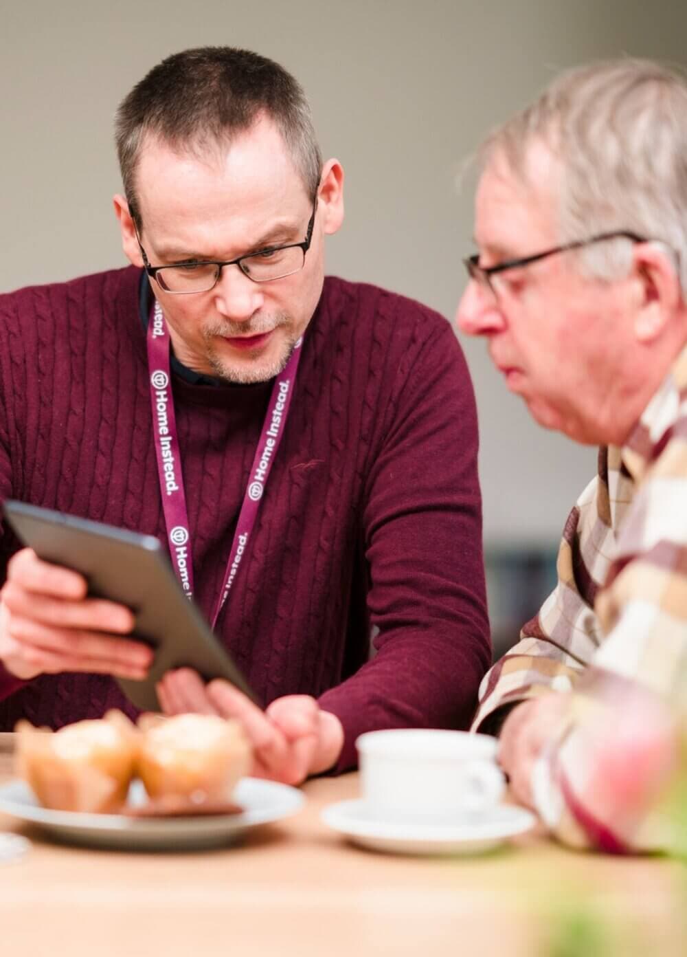 Two men sitting at a table, one showing the other a tablet; pastries and a coffee cup are also on the table. - Home Instead