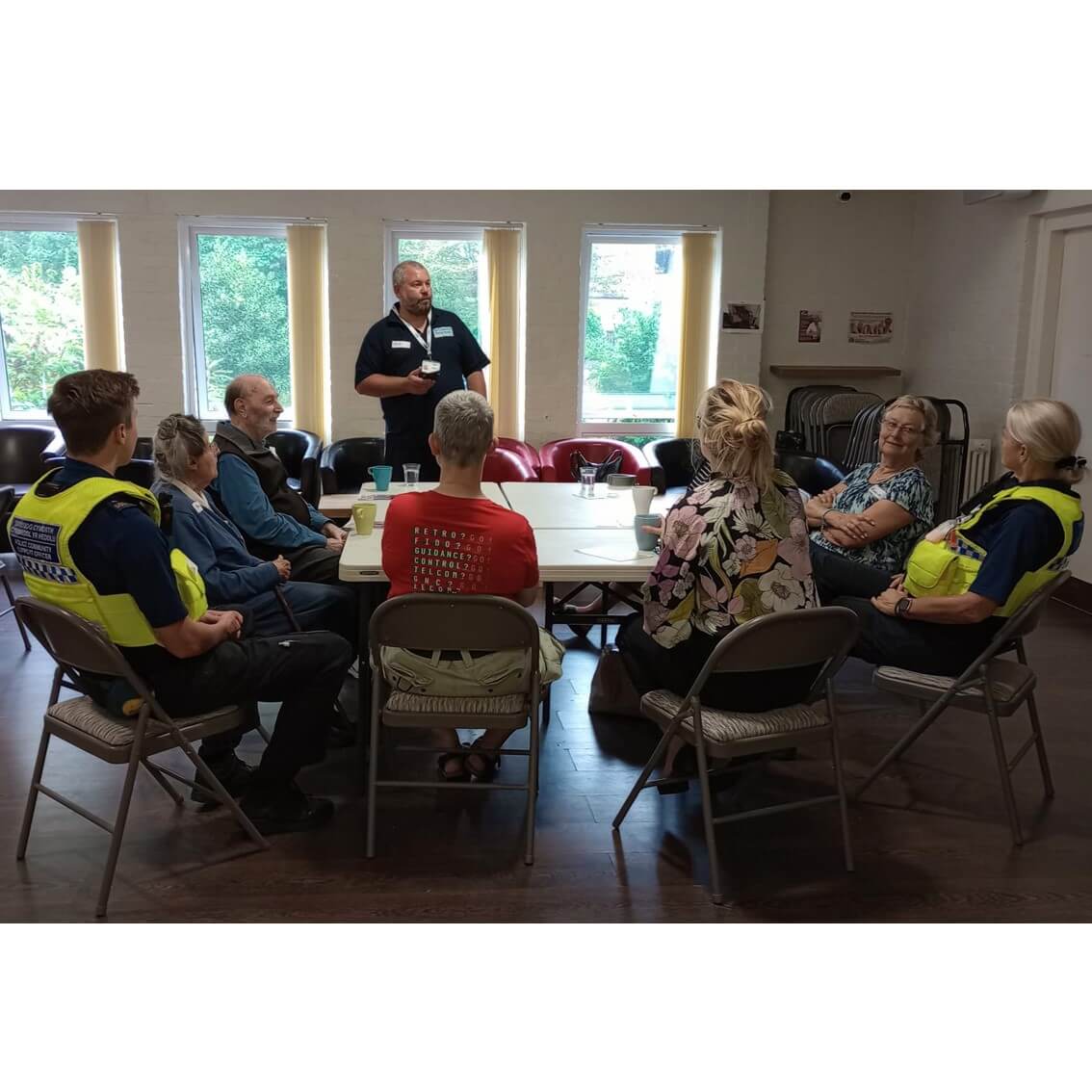 A group of people, some in uniform, are seated around a table in a room. One person is standing and speaking. - Home Instead