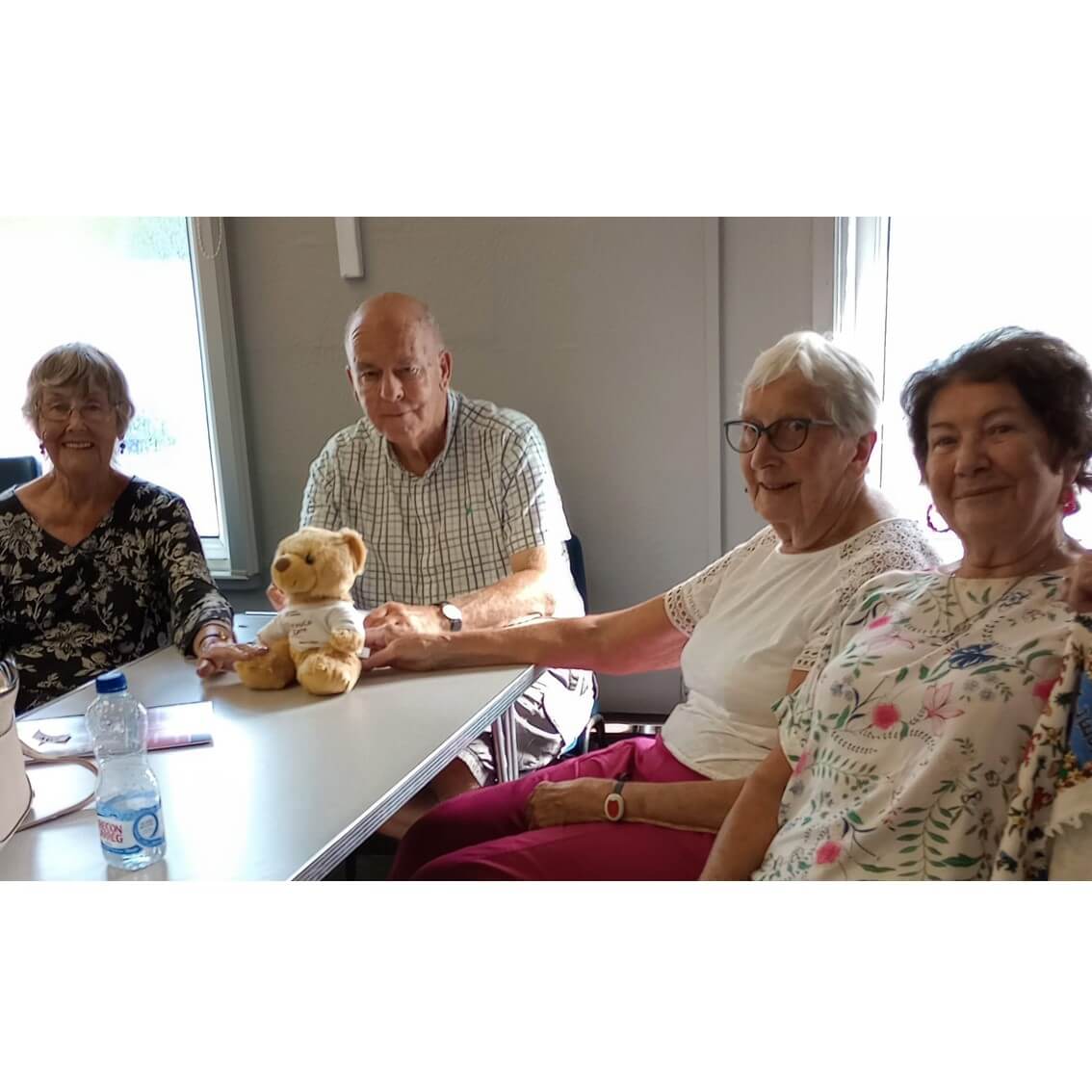 Four older adults sit around a table with a teddy bear, smiling. Water bottles and paperwork are on the table. - Home Instead
