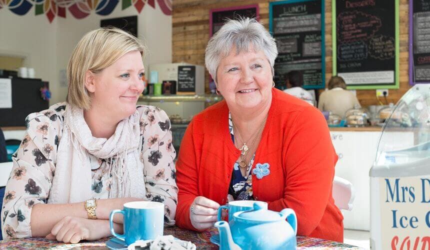 Two women smiling and sitting at a table with blue cups, a teapot, and menu boards in the background. - Home Instead