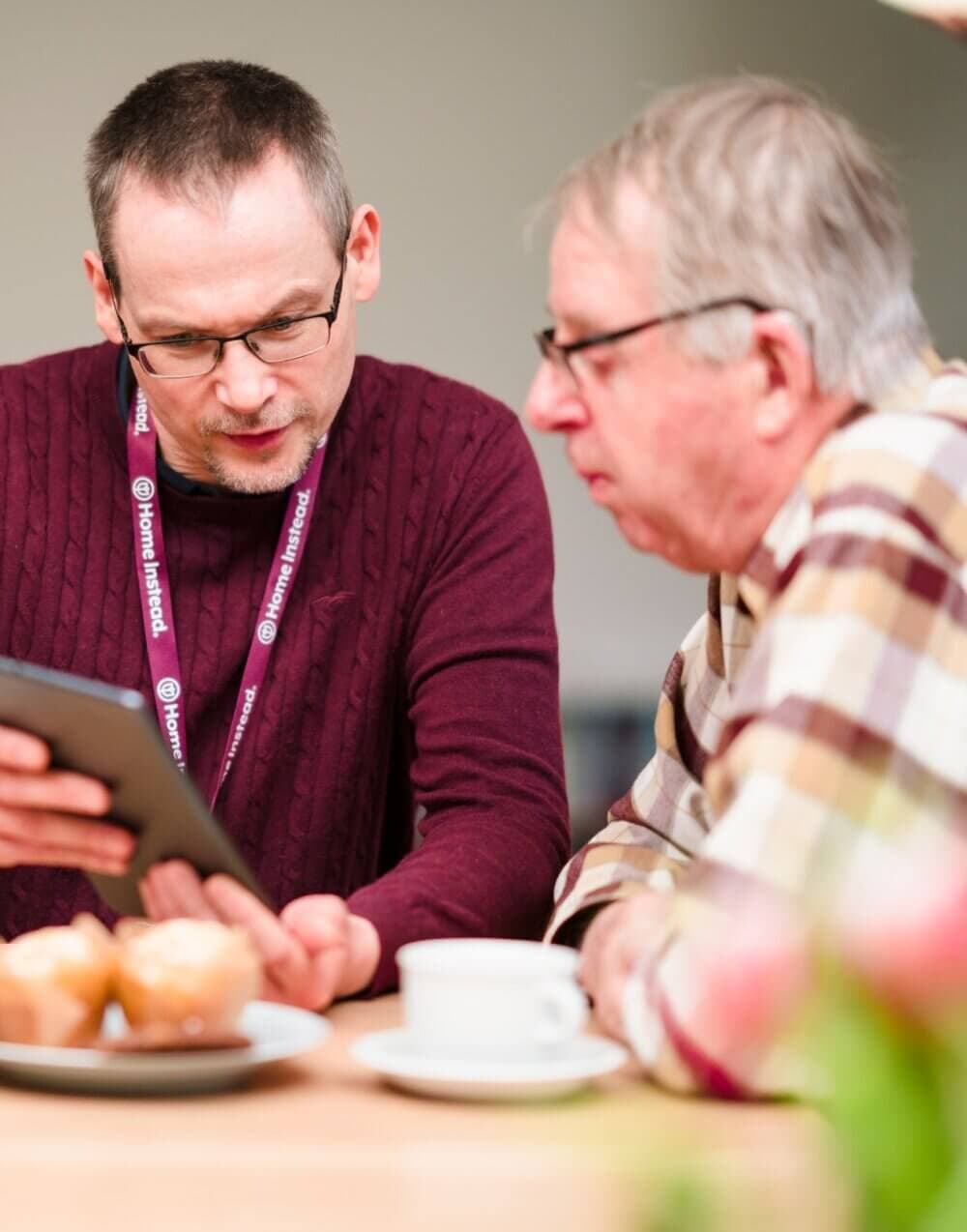 A Care Professional uses a tablet to assist an older man. They are sitting at a table with pastries and a cup of coffee. - Home Instead Poole