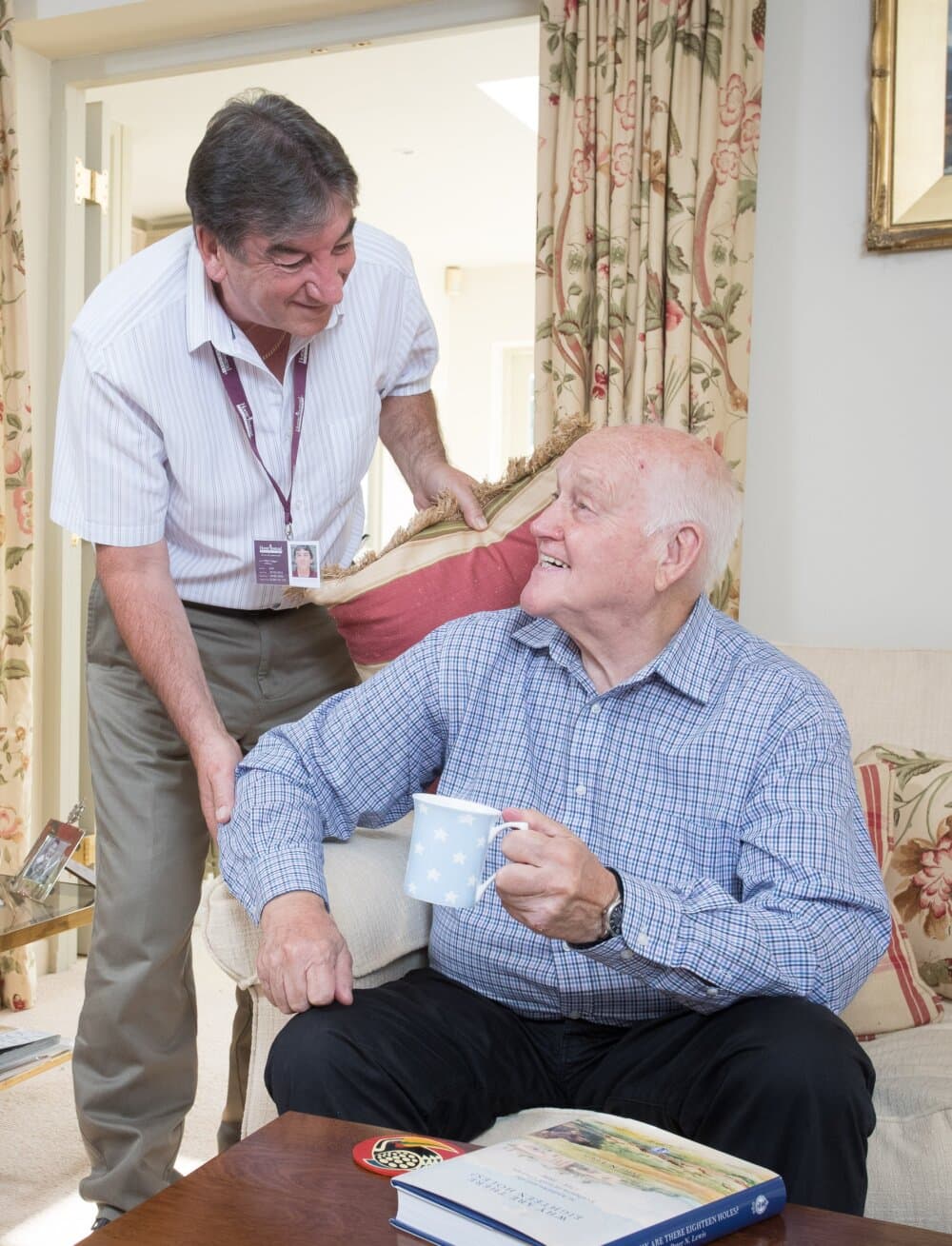 An elderly woman with white hair, glasses, and a blue cardigan smiles while holding a cookie on a couch. Plants are in the background. - Home Instead