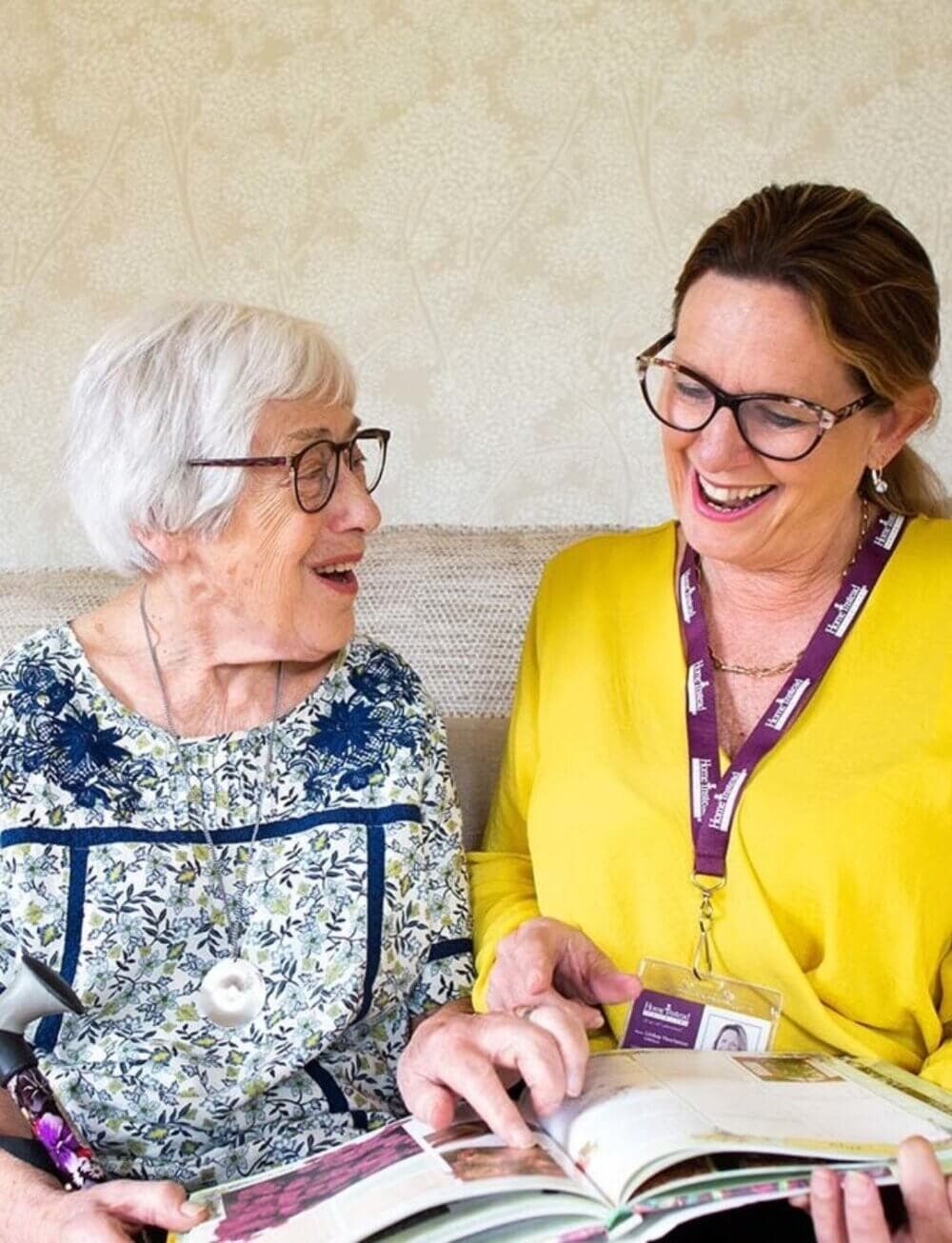 An elderly woman and a caregiver with glasses share a laugh while looking through a book together. - Home Instead