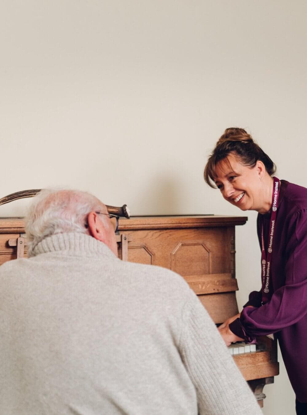 A woman smiles while playing the piano with an older man in a gray sweater, both enjoying their time together. - Home Instead