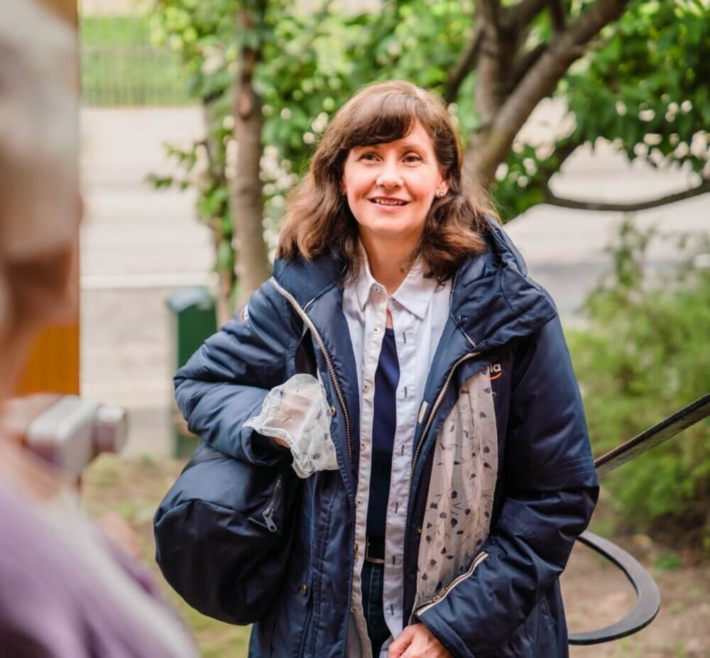 A woman outdoors wearing a blue jacket and white shirt, smiling, with greenery and a street in the background. - Home Instead