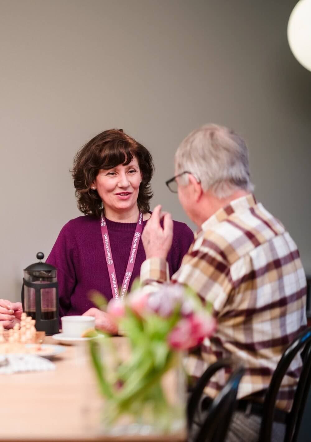Two people are talking at a table, one wearing a name tag. A French press and flowers are on the table. - Home Instead