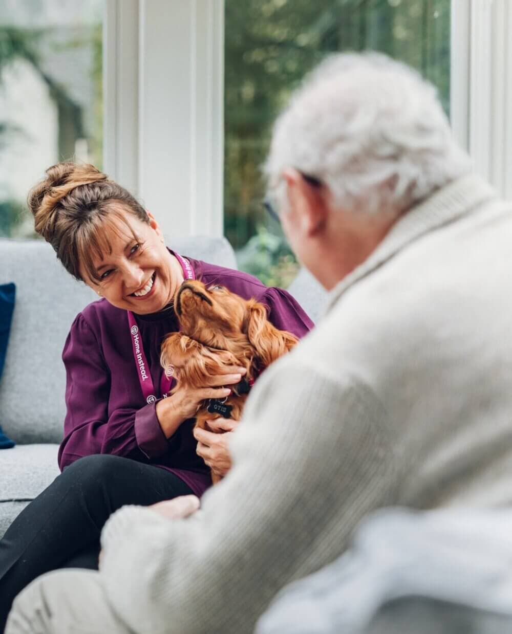 Woman smiling at elderly man while holding a dog, in a cosy room with large windows in the background. - Home Instead Poole