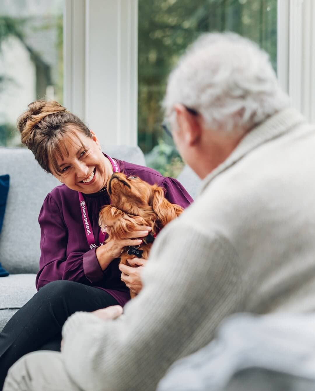 A woman holding a small dog smiles at an elderly man in a light sweater sitting on a couch indoors. - Home Instead Bournemouth & Christchurch