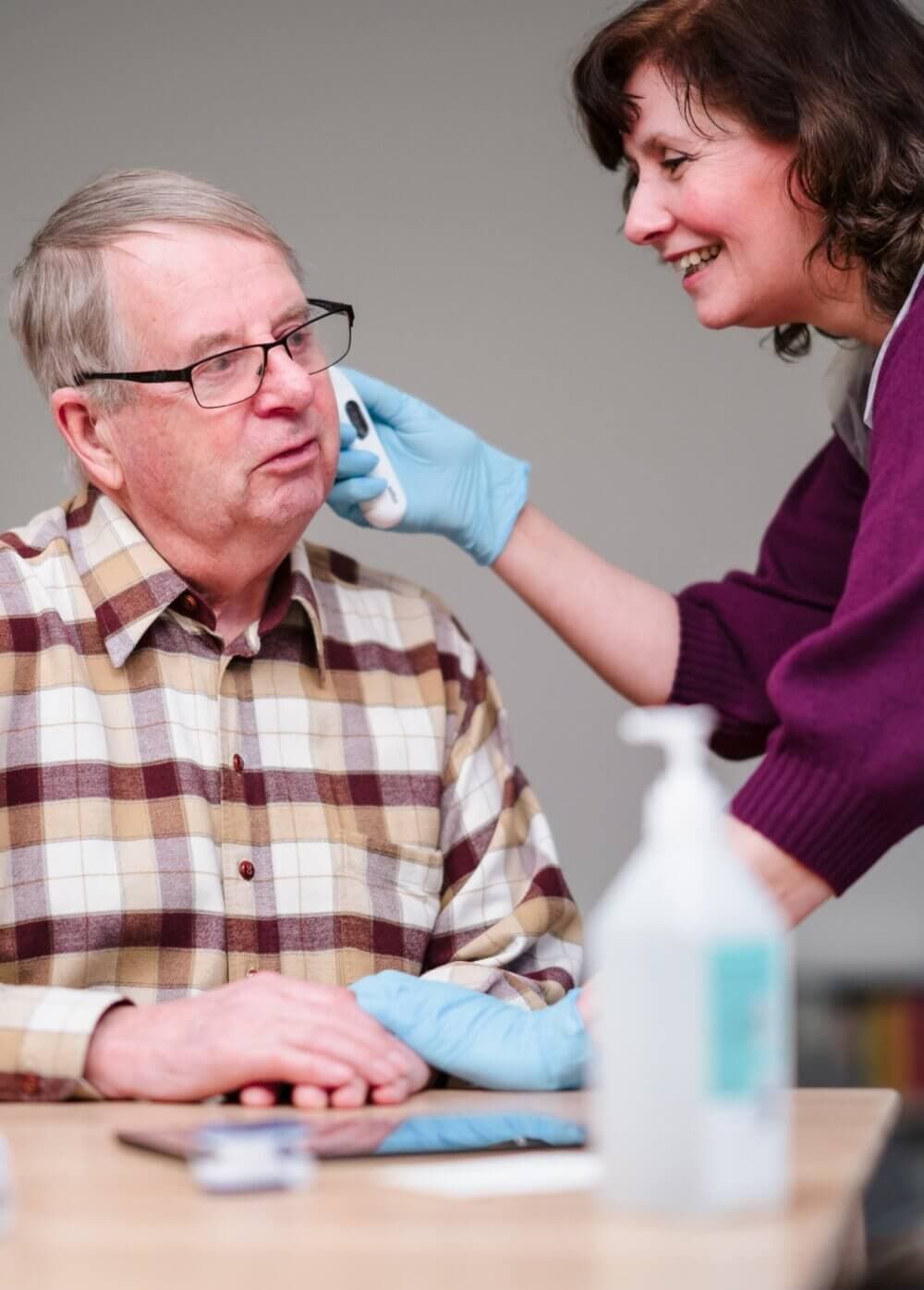 A healthcare worker wearing gloves and a mask checks the ear of an elderly man seated at a table. - Home Instead