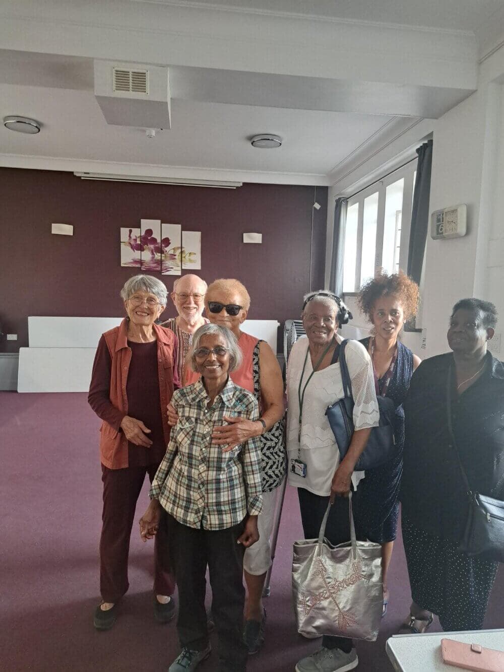 A group of seven smiling elderly individuals posing together in a room with purple walls and artwork. - Home Instead