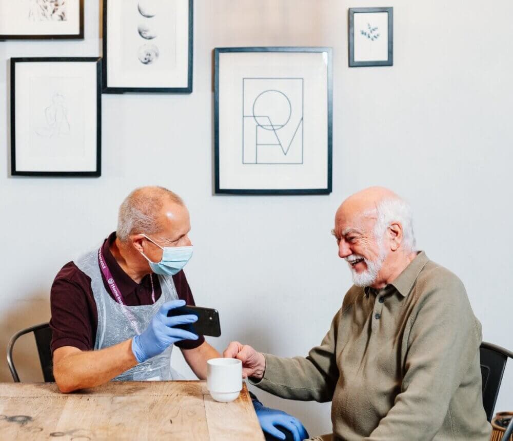 Two elderly men sit at a table. One shows something on a phone. Both are smiling. Framed pictures are on the wall. - Home Instead