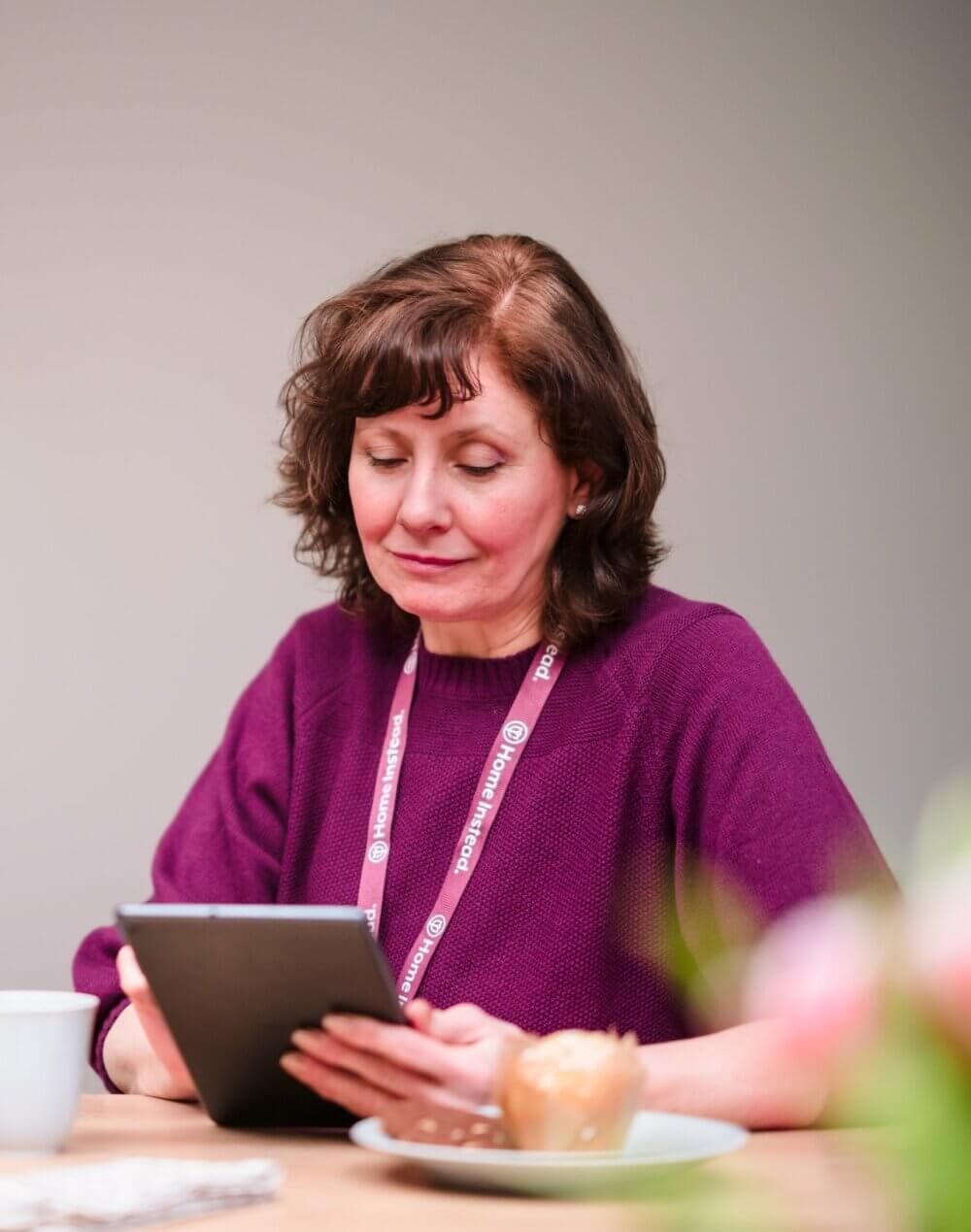 Woman with short brown hair, wearing a purple sweater, and a name badge, looking at a tablet, sitting at a table. - Home Instead