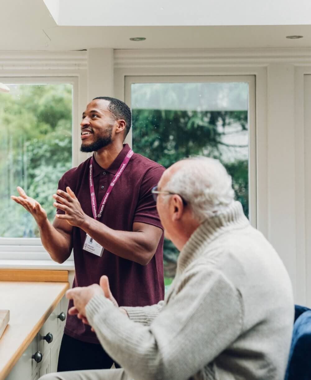 A man in a maroon shirt talks and gestures to an elderly man sitting at a table in a well-lit room. - Home Instead
