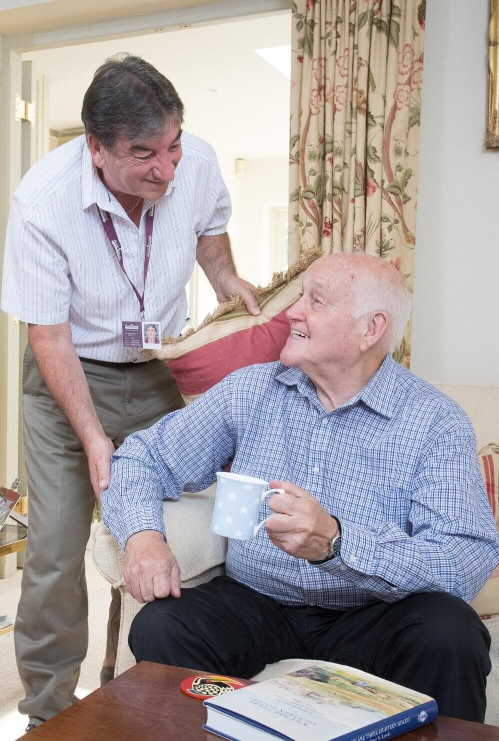 A Care Professional and an elderly man share a smile in a cosy living room. The elderly man holds a mug. - Home Instead Poole