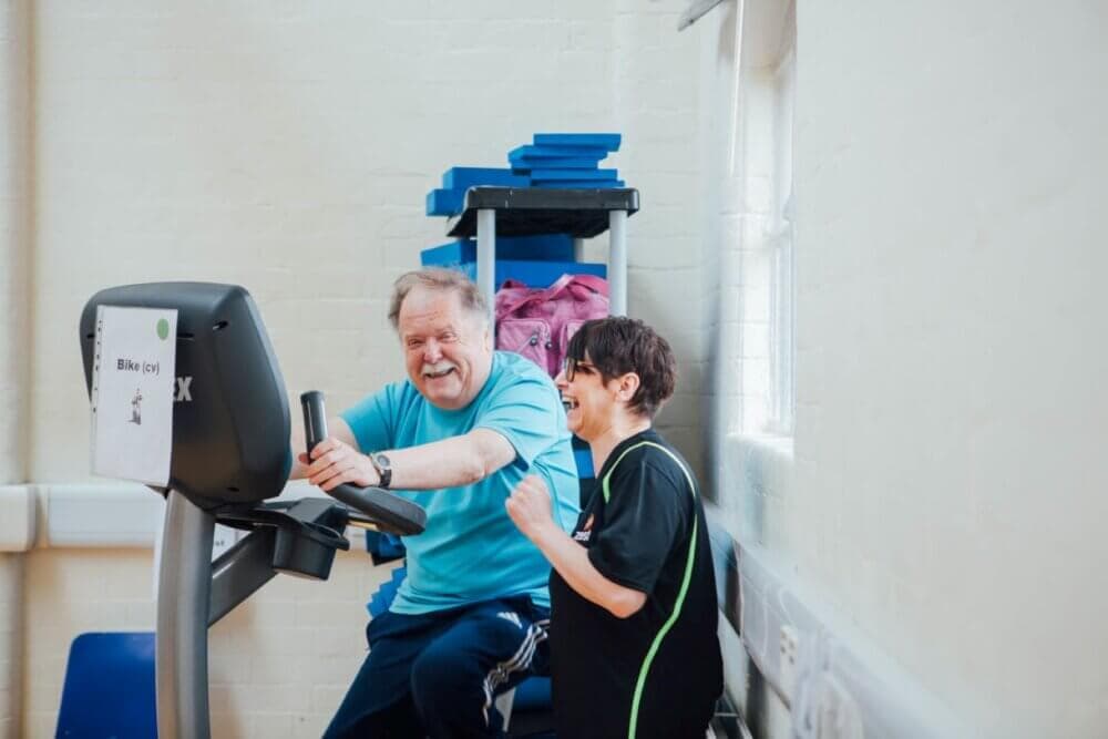 An elderly man exercises on a stationary bike while a woman in a tracksuit stands beside him, smiling and encouraging. - Home Instead