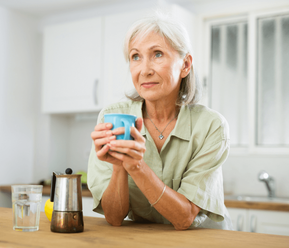 Elderly woman with gray hair holding a blue mug, standing in a kitchen with a pensive expression. - Home Instead