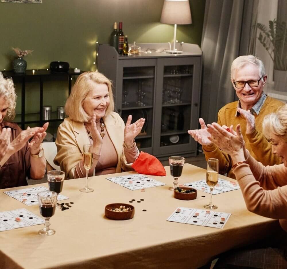 A group of elderly people sitting around a table, clapping and smiling, with drinks and game boards in front of them. - Home Instead