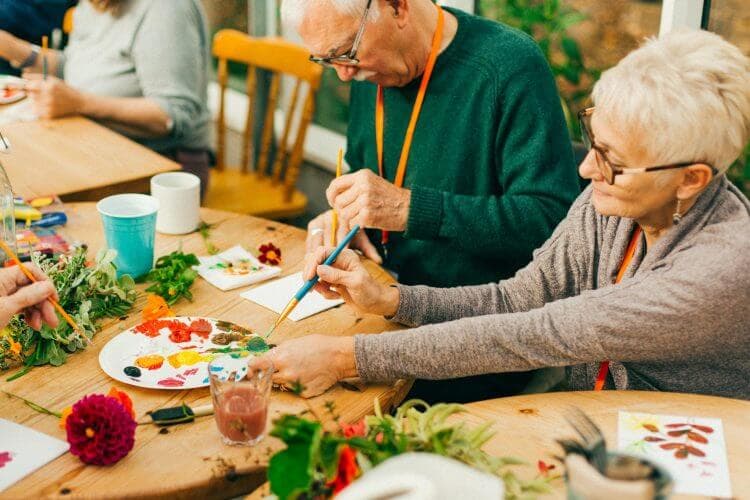 Two elderly people sitting at a table, painting and crafting together with various art supplies and plants. - Home Instead
