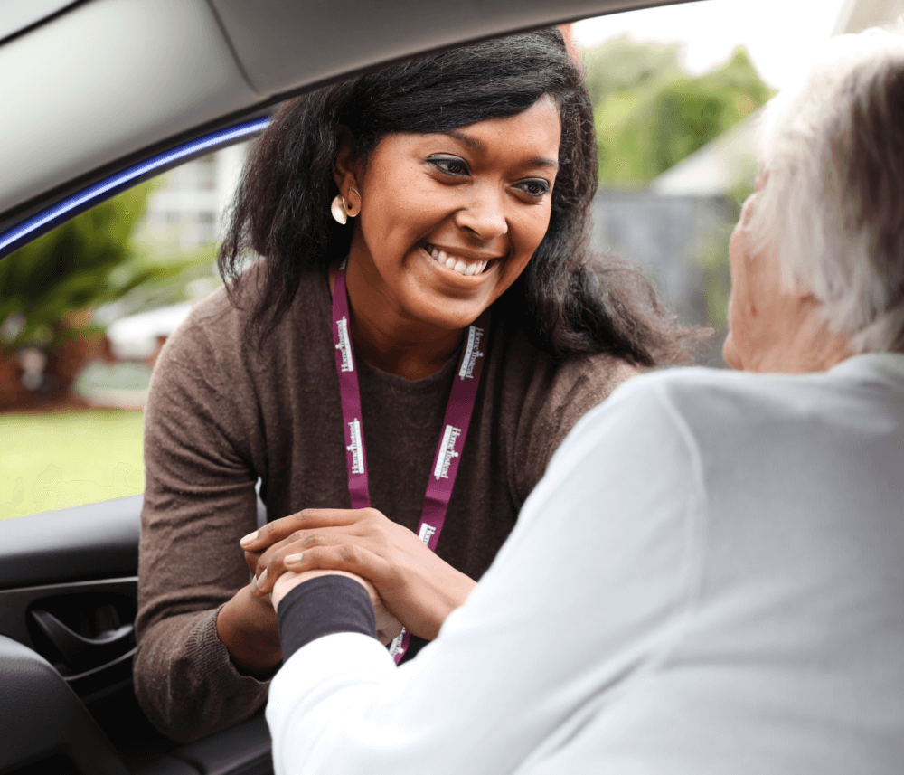 A smiling caregiver wearing a lanyard helps an older person in a car with a comforting hand. - Home Instead