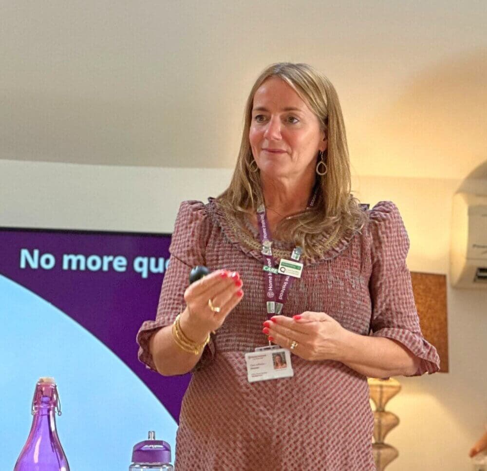 A woman in a purple dress is giving a presentation, holding a clicker with a screen in the background. - Home Instead
