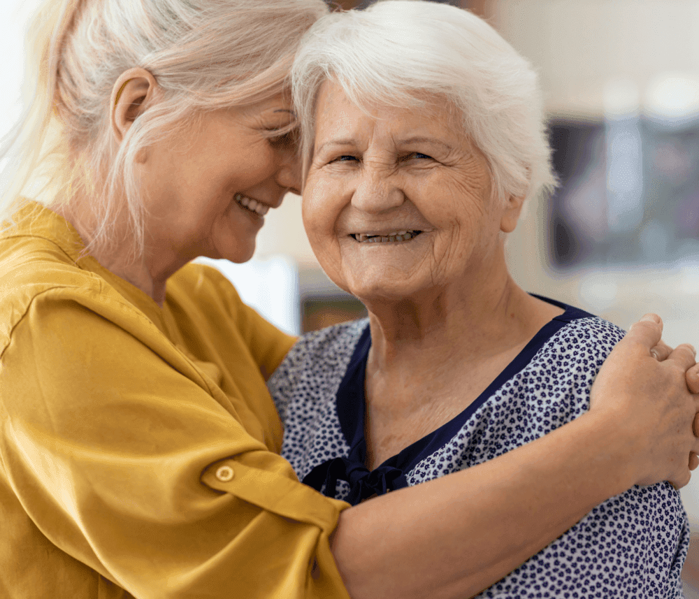 Two women smile and embrace affectionately, one is elderly with white hair, the other has blonde hair and wears a yellow shirt. - Home Instead